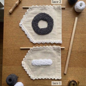 May include: Two beige fabric banners with lace trim, each with a different textured design in the center. One has a gray circle, the other a white rectangle. Wooden dowels, yarn balls, and a punch needle tool are also visible.