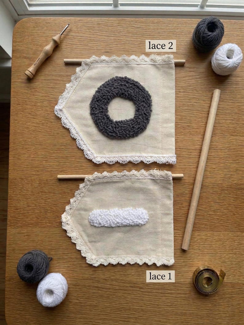 May include: Two beige fabric banners with lace trim, each with a different textured design in the center. One has a gray circle, the other a white rectangle. Wooden dowels, yarn balls, and a punch needle tool are also visible.