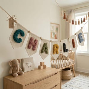 May include: A nursery banner spelling "CHARLIE" with colorful, textured letters on off-white pennants. The banner hangs above a wooden dresser with a crochet elephant, a book, and wooden blocks. A crib and basket are in the background.