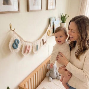 May include: A nursery scene with a baby and a woman. A banner spelling "SHILO" hangs above a wooden crib. The room is decorated with framed animal prints and a stuffed elephant. The baby is wearing a cream-colored sweater.