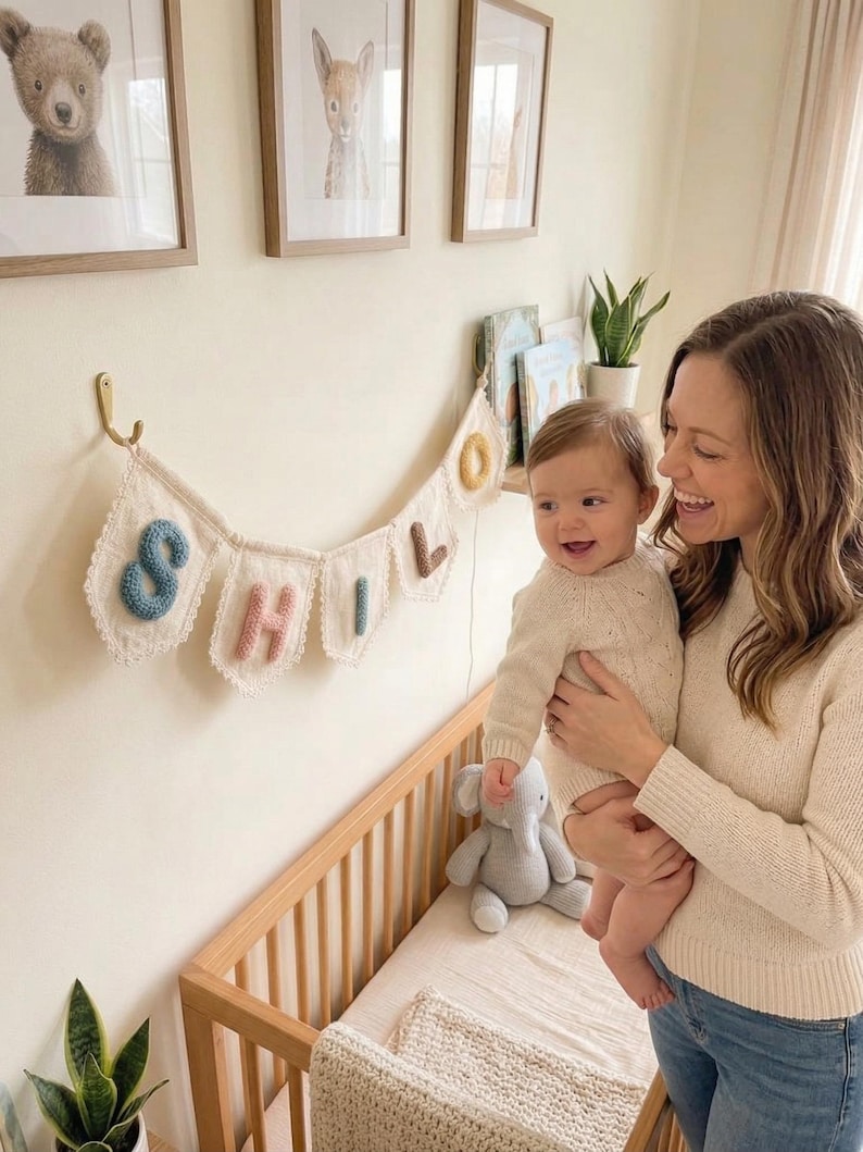 May include: A nursery scene with a baby and a woman. A banner spelling "SHILO" hangs above a wooden crib. The room is decorated with framed animal prints and a stuffed elephant. The baby is wearing a cream-colored sweater.