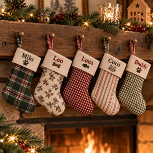 Könnte beinhalten: Fünf Weihnachtsstrümpfe aus Stoff mit cremefarbenen Bündchen, jeweils personalisiert mit einem Namen und einem kleinen Symbol. Die Strümpfe zeigen verschiedene Muster, darunter grünes Schottenkaro, Stechpalmen-Druck, rotes Vichy-Karo, Streifen und grünes Vichy-Karo.