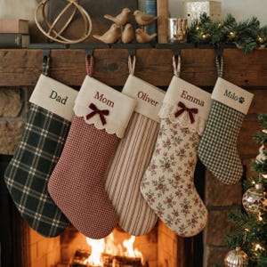May include: Five hanging Christmas stockings with personalized names. The first is dark green plaid, the second is red gingham with a bow, the third has red vertical stripes, the fourth features a floral print with lace and a bow, and the fifth is green gingham.