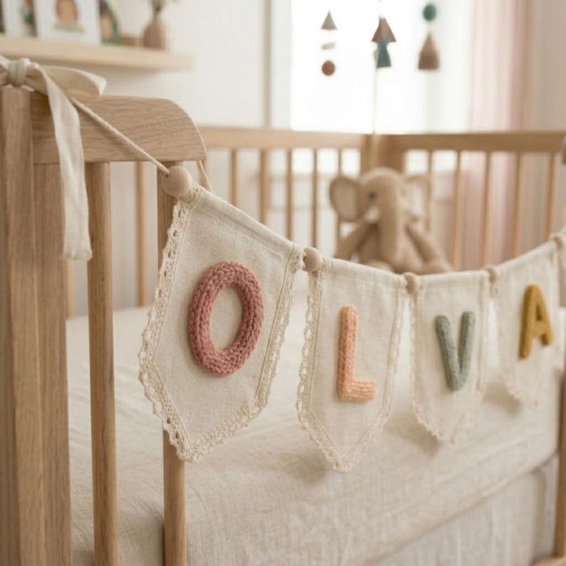 May include: A banner spelling "OLIVA" hangs on a wooden crib. Each letter is a different color: pink, peach, green, and yellow. The banner is made of off-white fabric with a lace trim and wooden beads.