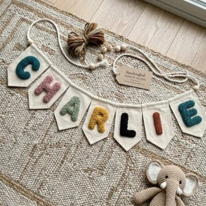 May include: A personalized nursery banner spelling "CHARLIE" in colorful, textured letters. The banner is made of off-white fabric pennants, strung together with a white rope. A tag reads "Handcrafted Nursery Wilder".