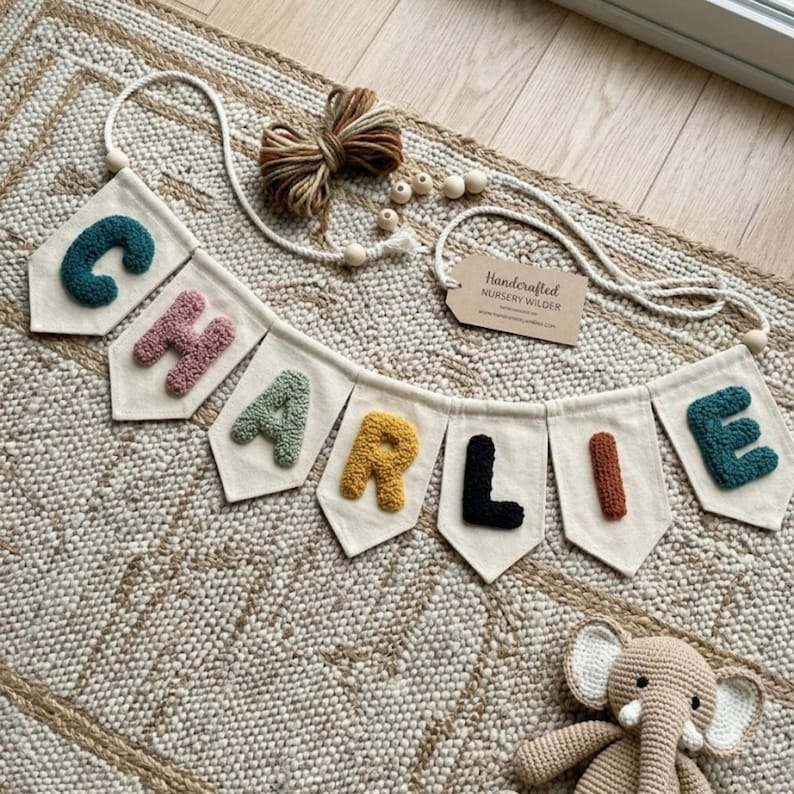 May include: A personalized nursery banner spelling "CHARLIE" in colorful, textured letters. The banner is made of off-white fabric pennants, strung together with a white rope. A tag reads "Handcrafted Nursery Wilder".