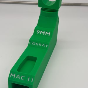 May include: A bright green, 3D-printed object with the text "9MM", "COBRAY", and "MAC II" in white. The object has a rectangular base with a hollowed-out section and a curved top. The object is likely a tool or accessory.