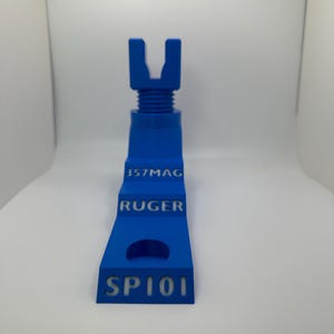 May include: A blue 3D-printed object with the text "357MAG", "RUGER", and "SP101". The object has a U-shaped top and a stepped design. The object is a gun accessory.