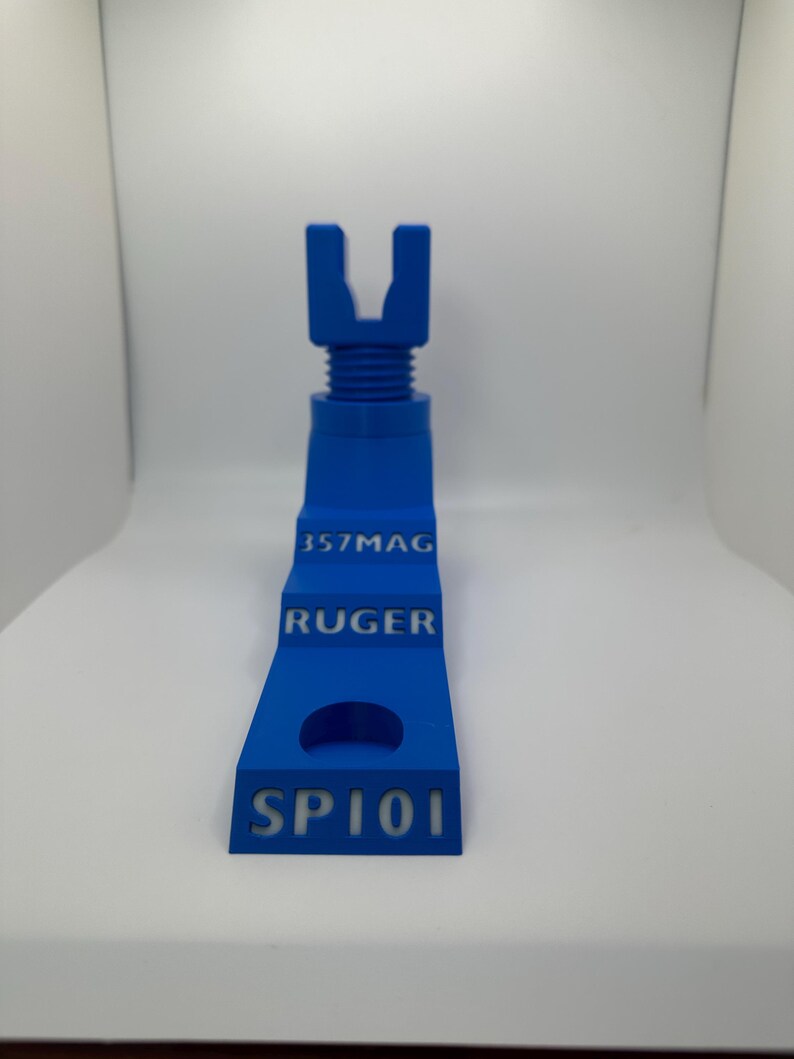 May include: A blue 3D-printed object with the text "357MAG", "RUGER", and "SP101". The object has a U-shaped top and a stepped design. The object is a gun accessory.