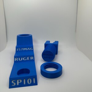 May include: Blue 3D-printed accessories, including a stepped stand with "357MAG", "RUGER", and "SP101" text, a cylindrical piece with "RUGER SP101", and a circular threaded component. The items are set against a white background.