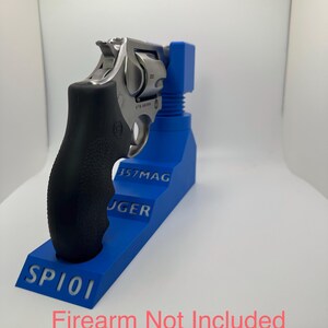 May include: A stainless steel revolver with a black grip is displayed on a blue stand. The stand has the text "357MAG", "RUGER", and "SP101". The text "Firearm Not Included" is at the bottom.