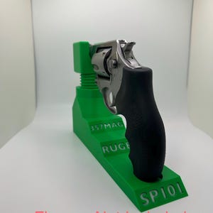 May include: A stainless steel revolver with a black grip rests on a bright green stand. The stand has the text "357MAG", "RUGE", and "SP101" printed on it. The text "Firearm Not Included" is printed in pink.