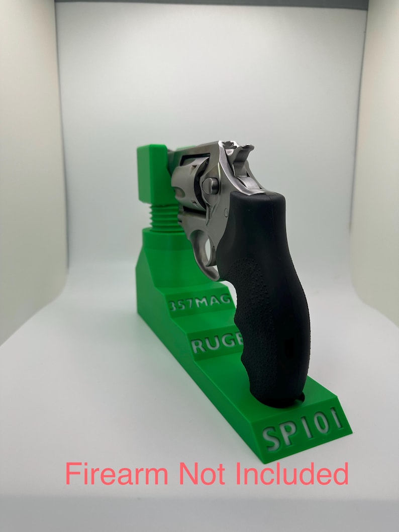 May include: A stainless steel revolver with a black grip rests on a bright green stand. The stand has the text "357MAG", "RUGE", and "SP101" printed on it. The text "Firearm Not Included" is printed in pink.