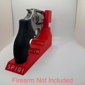 May include: A stainless steel revolver with a black grip is displayed on a bright red stand. The stand has the text "SP101", "357MAG", and "RUGER" in white. The image also includes the text "Firearm Not Included" in pink.