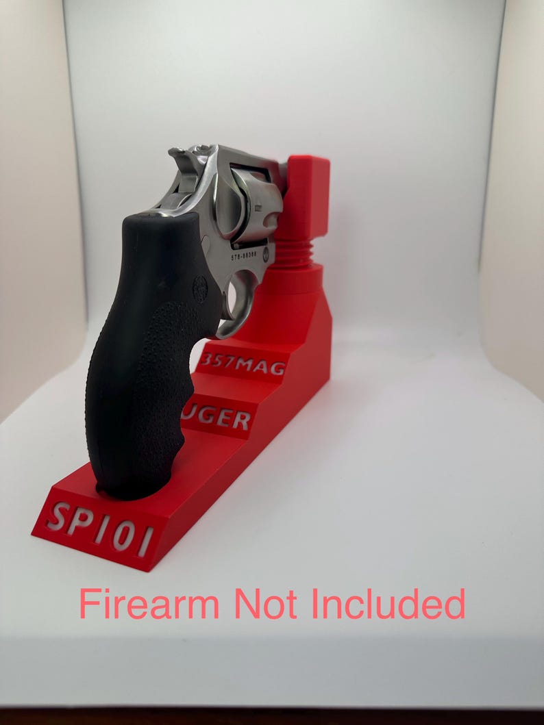 May include: A stainless steel revolver with a black grip is displayed on a bright red stand. The stand has the text "SP101", "357MAG", and "RUGER" in white. The image also includes the text "Firearm Not Included" in pink.