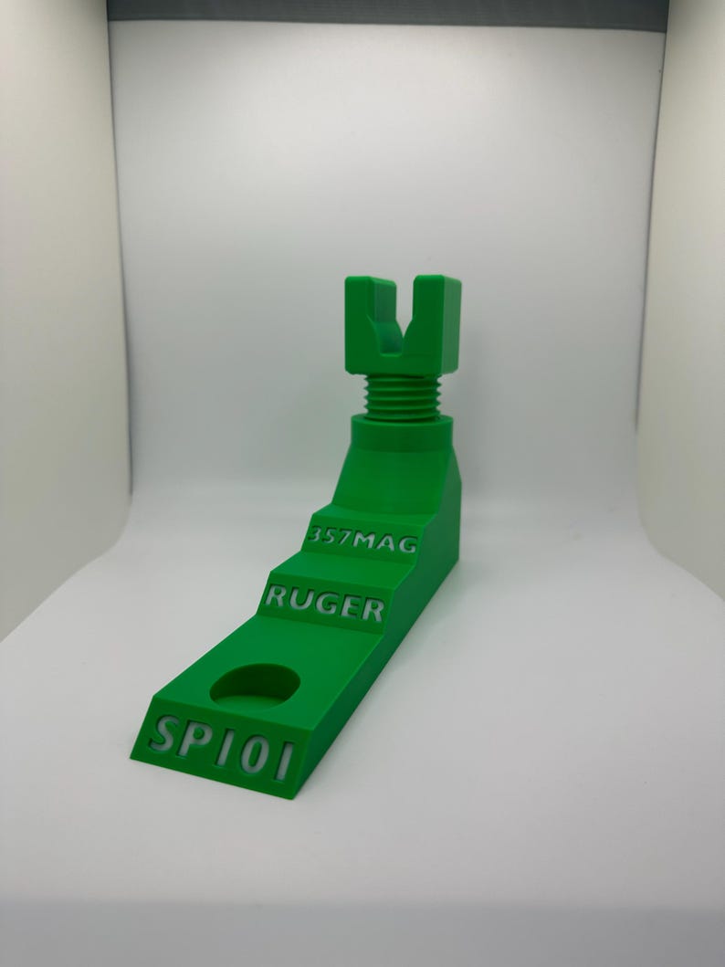 May include: A bright green, 3D-printed gun stand with the text "SP101", "RUGER", and "357MAG". The stand has a stepped design with a circular hole and a U-shaped top. The stand is designed to hold a gun.