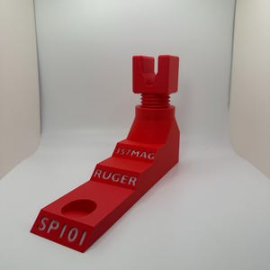 May include: A bright red gun stand with the text "SP101", "RUGER", and "357MAG" etched into the side. The stand has a stepped design and a U-shaped top, likely for holding a firearm. The stand is made of a solid material.