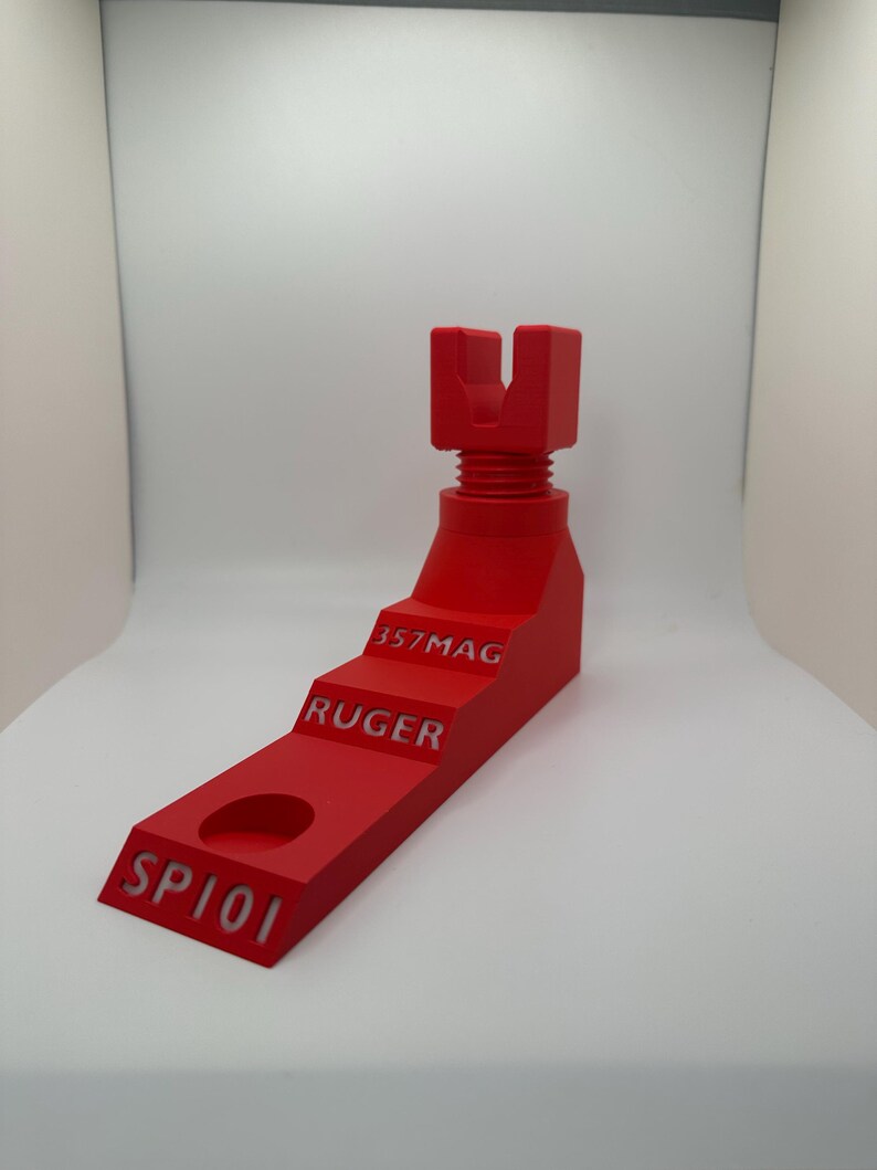 May include: A bright red gun stand with the text "SP101", "RUGER", and "357MAG" etched into the side. The stand has a stepped design and a U-shaped top, likely for holding a firearm. The stand is made of a solid material.