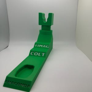 Puede incluir: Un soporte verde brillante impreso en 3D con el texto "ANACONDA", "COLT" y "44MAG". El soporte tiene una parte superior en forma de U y una base rectangular con un recorte curvo. El soporte está diseñado para sostener un objeto.