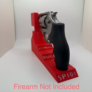 May include: A red 3D-printed firearm stand with the text "357MAG", "RUGER", and "SP101". A silver revolver with a black grip rests on the stand. The text "Firearm Not Included" is visible.