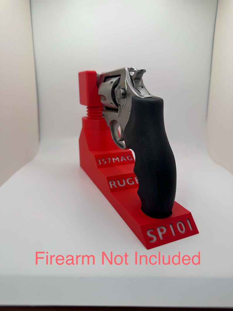 May include: A red 3D-printed firearm stand with the text "357MAG", "RUGER", and "SP101". A silver revolver with a black grip rests on the stand. The text "Firearm Not Included" is visible.