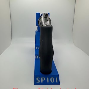 May include: A blue 3D-printed firearm stand with the text "SP101" and "35 R" on the side. A black-handled revolver is placed in the stand. The text "Firearm Not Included" is at the bottom.