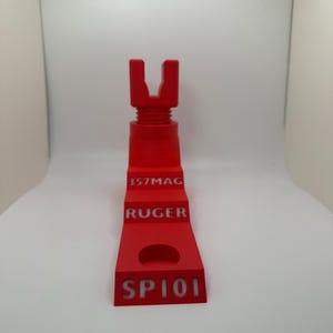 May include: A bright red 3D-printed object with the text "357MAG", "RUGER", and "SP101". The object has a U-shaped top and a stepped design with a circular cutout. The item is designed for a firearm.