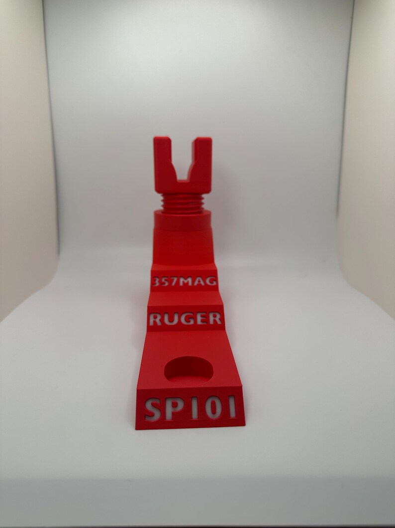 May include: A bright red 3D-printed object with the text "357MAG", "RUGER", and "SP101". The object has a U-shaped top and a stepped design with a circular cutout. The item is designed for a firearm.