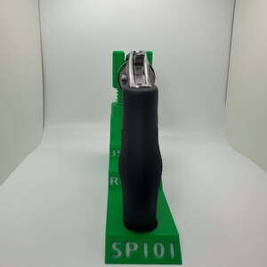 May include: A black handgun with a silver cylinder is held in a bright green stand. The stand has the text "SP101" and "Firearm Not Included" in pink. The stand is designed to hold the handgun.