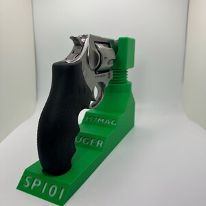 May include: A stainless steel revolver with a black grip rests on a bright green stand. The stand is labeled "SP101", "RUGER", and "357MAG". The text "Firearm Not Included" is visible in red.