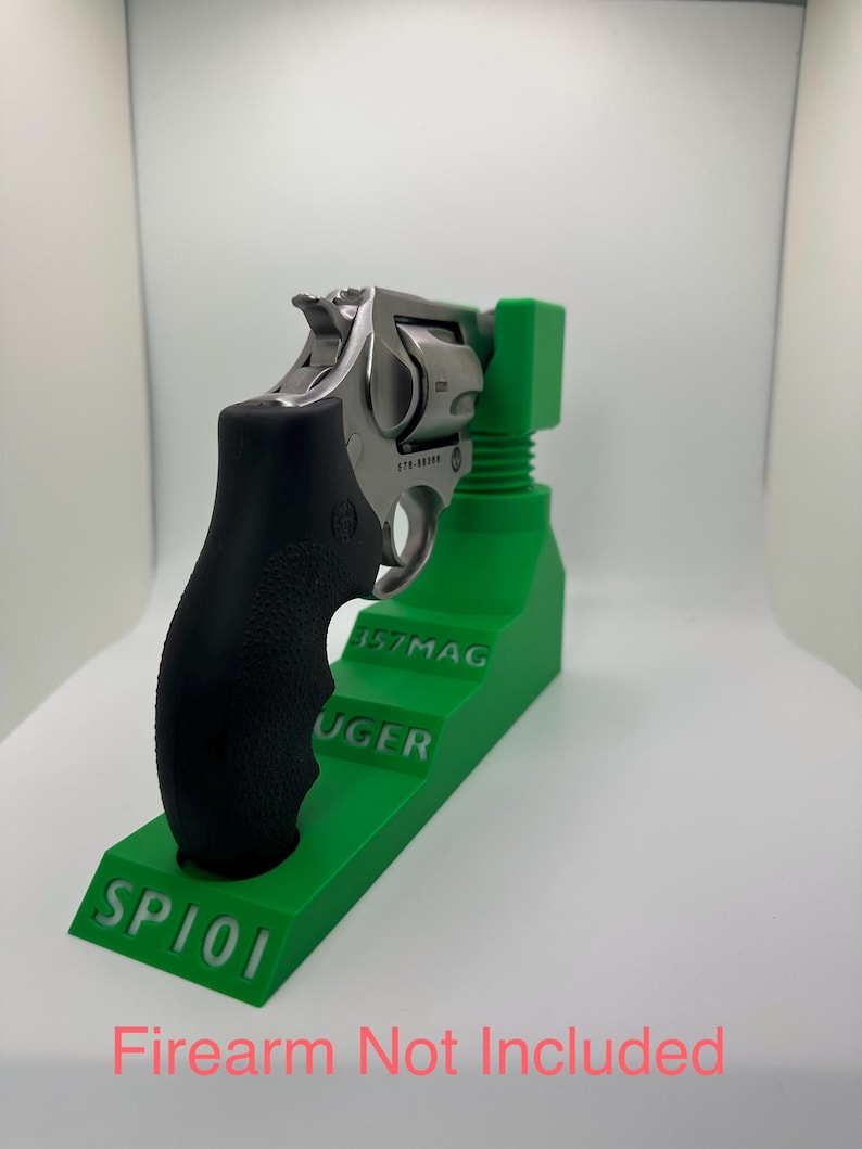 May include: A stainless steel revolver with a black grip rests on a bright green stand. The stand is labeled "SP101", "RUGER", and "357MAG". The text "Firearm Not Included" is visible in red.