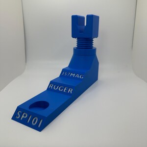 May include: A vibrant blue 3D-printed object with the text "SP101", "RUGER", and "357MAG". The object has a stepped design with a hole and a threaded section. The top features a U-shaped component.