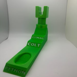 May include: A bright green 3D-printed stand for a Colt Python firearm. The stand features the text "357 MAG", "COLT", and "PYTHON". The stand has a slot for the firearm and a threaded post.