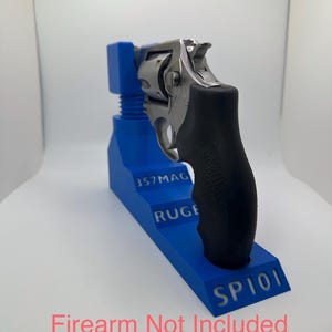 May include: A blue firearm stand with the text "357MAG RUGE SP101". A silver and black handgun rests on the stand. The text "Firearm Not Included" is at the bottom.