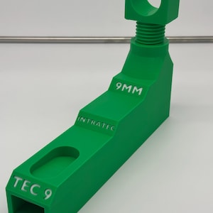 May include: A bright green, 3D-printed object with the text "TEC 9", "INTRATEC", and "9MM". The object has a rectangular base with a curved cutout, an angled midsection, and a threaded top with a U-shaped cradle.