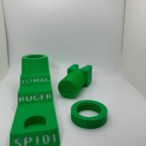 May include: Bright green 3D-printed firearm parts, including a stepped component with "357MAG", "RUGER", and "SP101" text, a cylindrical piece with "RUGER SP101", and a circular ring. The parts are displayed on a white surface.