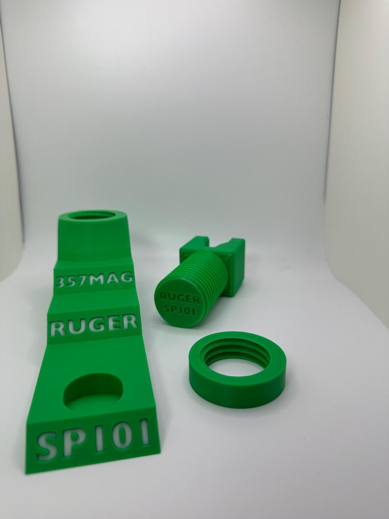 May include: Bright green 3D-printed firearm parts, including a stepped component with "357MAG", "RUGER", and "SP101" text, a cylindrical piece with "RUGER SP101", and a circular ring. The parts are displayed on a white surface.