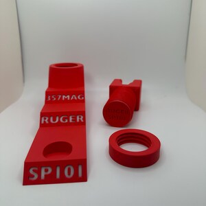 May include: A set of four bright red 3D-printed accessories. The set includes a stepped stand with "357MAG", "RUGER", and "SP101" text, a threaded cylinder with "RUGER SP101" text, and a ring.
