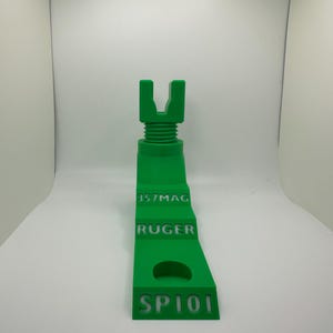 May include: A bright green 3D-printed object with the text "357MAG", "RUGER", and "SP101". The object has a U-shaped top and a threaded section. It is designed to hold a firearm.