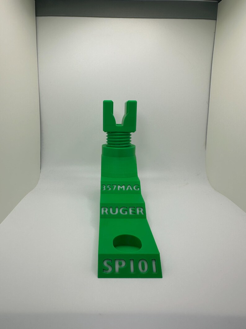 May include: A bright green 3D-printed object with the text "357MAG", "RUGER", and "SP101". The object has a U-shaped top and a threaded section. It is designed to hold a firearm.