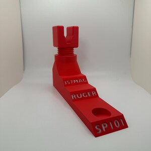 May include: A bright red, 3D-printed object with a stepped design. The object has a threaded top section and text that reads "357MAG", "RUGER", and "SP101". It features a circular hole near the base.
