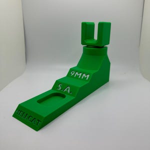 May include: A bright green 3D-printed object with the text "9MM", "S.A.", and "HELLCAT". The object has a stepped design with a U-shaped top. The object is designed to hold a firearm.