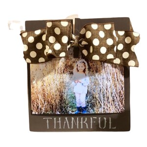 May include: A brown wooden picture frame with a white polka dot bow. The frame has a photo of a young girl in a field and the word "THANKFUL" written on the bottom.