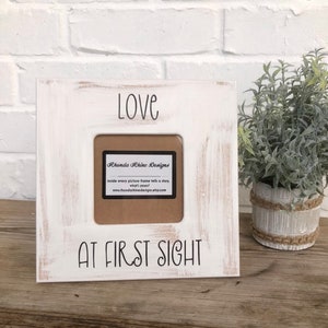 May include: White picture frame with distressed paint and the text "Love at First Sight". The frame has a small opening for a photo and a brown paper label with the text "Rhonda Rhine Designs" and a website address.