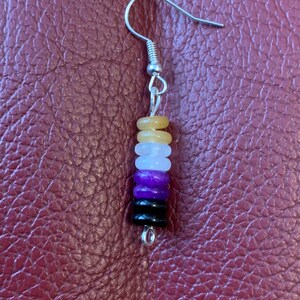 May include: A single earring with a silver hook and a stack of colourful beads. The beads are in the colours of the non-binary pride flag: yellow, white, purple, black, and a light yellow. The earring is displayed against a textured, reddish-brown surface.