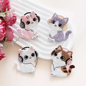 Puede incluir: Cuatro parches bordados con gatos de dibujos animados que llevan auriculares. Un gato es rosa y blanco, otro es morado y blanco, otro es blanco sólido y otro es marrón, blanco y tostado. Cada gato tiene ojos grandes y una nariz pequeña.