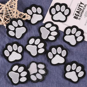 May include: A collection of ten embroidered paw print patches, each featuring a black border and white thread detailing for the pads and toes. The patches are arranged in a scattered pattern, with one patch showing its white adhesive backing.