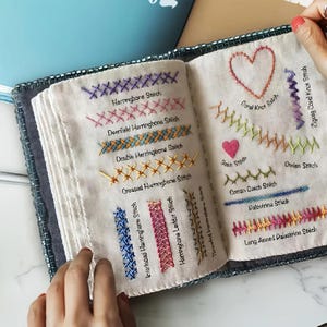 May include: A fabric embroidery sampler book featuring various hand-stitched patterns on off-white pages. Visible stitches include Herringbone, Deerfield, Double, Crossed, Interlaced, Ladder, Threaded, Coral Knot, Zigzag, Satin, Cretan, Catch, Palestrina, and Long Armed Palestrina.