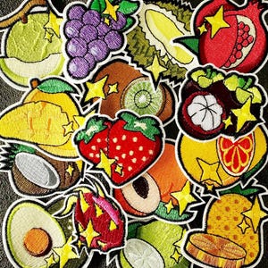 May include: A collection of embroidered patches featuring various fruits including strawberries, grapes, pomegranates, mangos, pineapples, avocados, and dragon fruit, each accented with small yellow star-shaped highlights and white outlines.