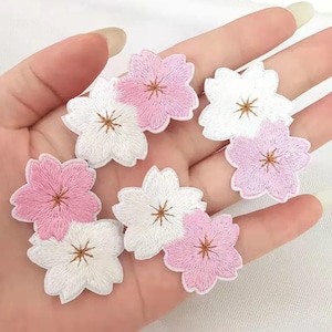 May include: A collection of six embroidered flower patches in shades of light pink and white. Each patch features a five-petaled blossom design with a small, brown, star-shaped center detail, showcasing intricate thread work and defined edges.